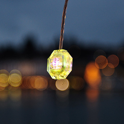 Diamante pendant light: small faceted crystal-like geometric lamp glowing yellow-green, suspended on a wire against a dusk bokeh background