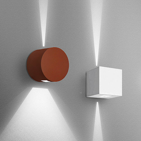 Two Artemide Effetto wall sconces: cylindrical rust-red and cubic white, both projecting dramatic upward and downward light beams on gray wall