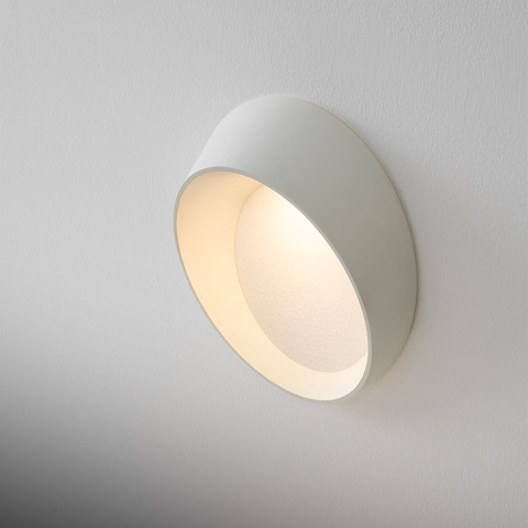 Artemide Cleomede white wall lamp with circular tilted shade emitting warm diffused light against a white wall