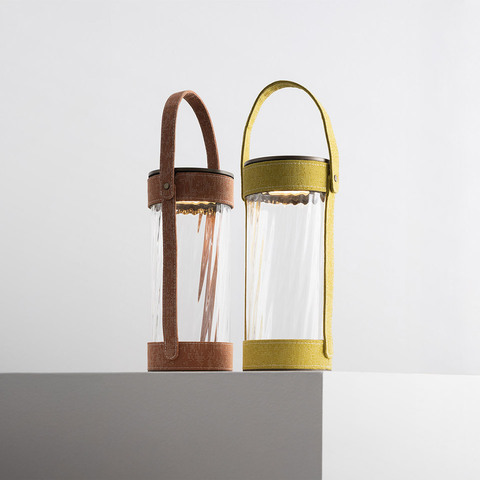 Two Artemide Laya portable lanterns with cylindrical glass body and fabric handle: left brown leather, right olive green