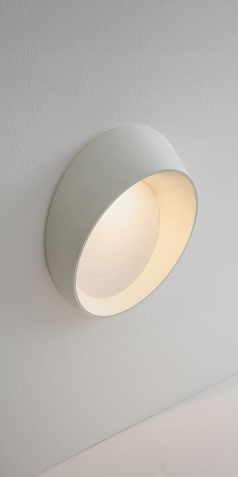Artemide Cleomede wall lamp: white circular disc-shaped fixture with warm glowing interior, mounted flush on white wall
