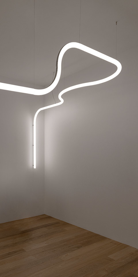 Artemide Alphabet of Light: glowing white LED tube bent into an abstract suspended shape, with a vertical drop element, against white walls