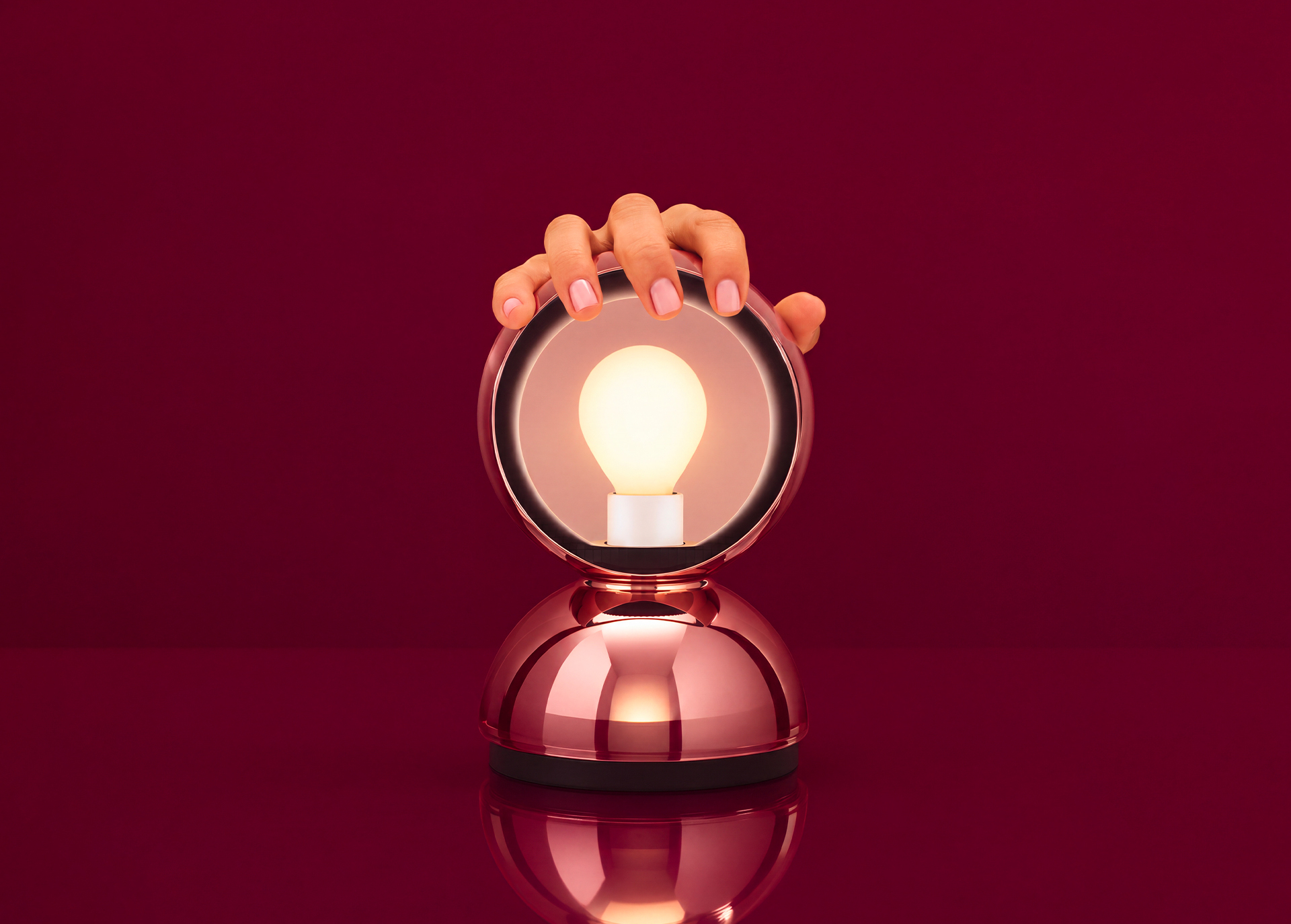 Hand touching a rose gold table lamp with circular mirror frame and glowing Edison bulb on a reflective surface, red background