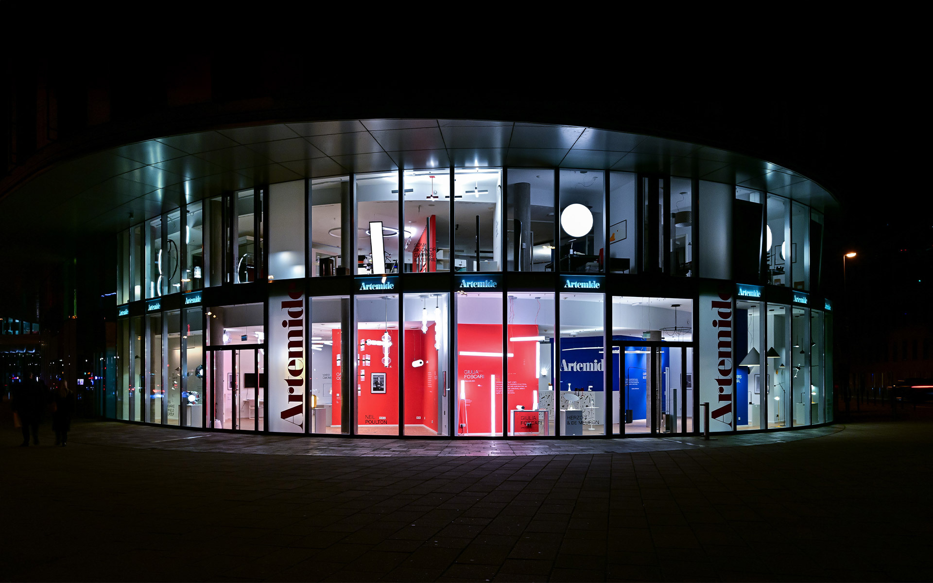 Artemide flagship store exterior at night, curved glass facade illuminated with red and blue display areas showcasing lighting collections
