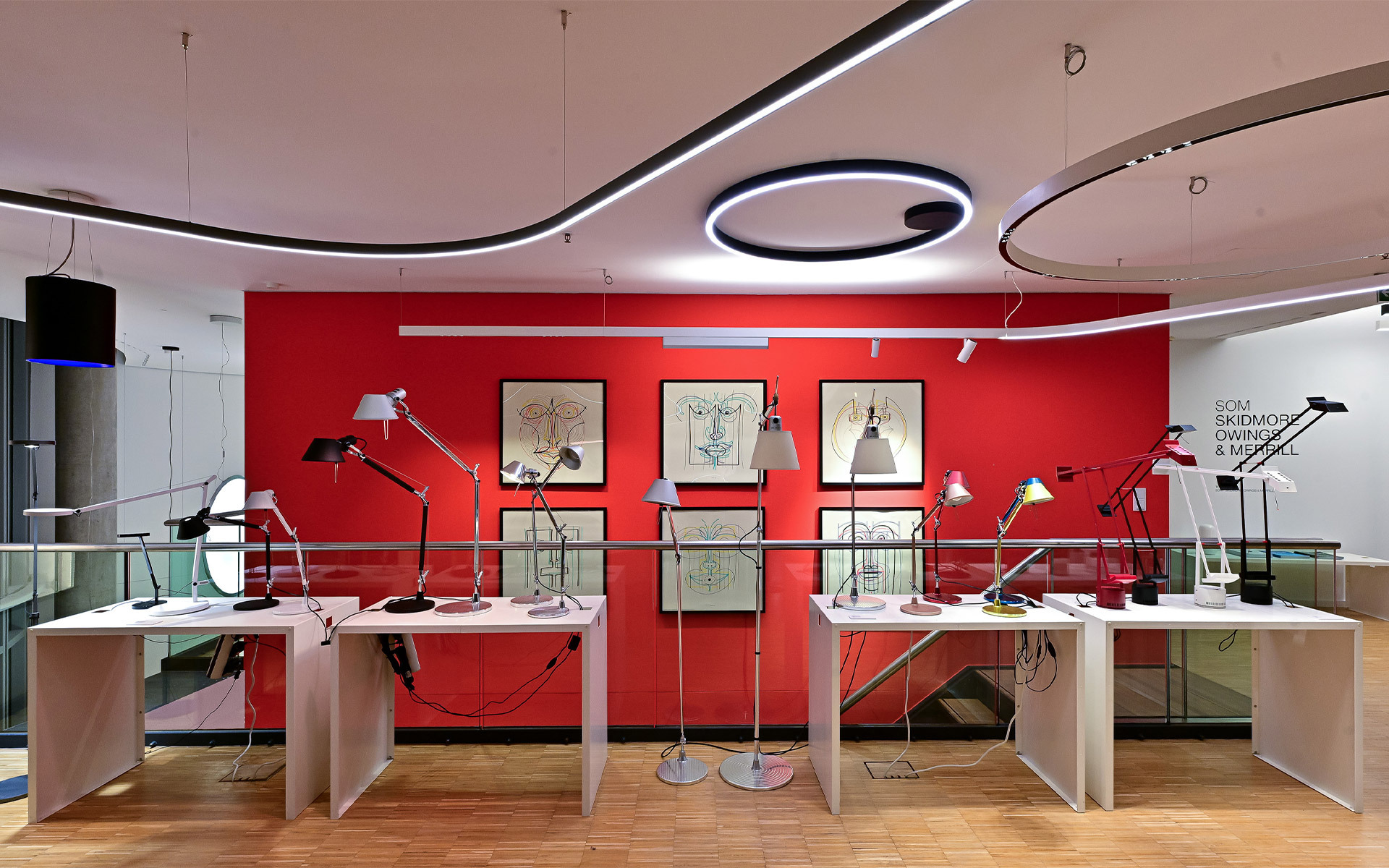 Artemide showroom displaying multiple Tolomeo desk and floor lamps on white tables against a bold red wall, with circular LED ceiling lights.