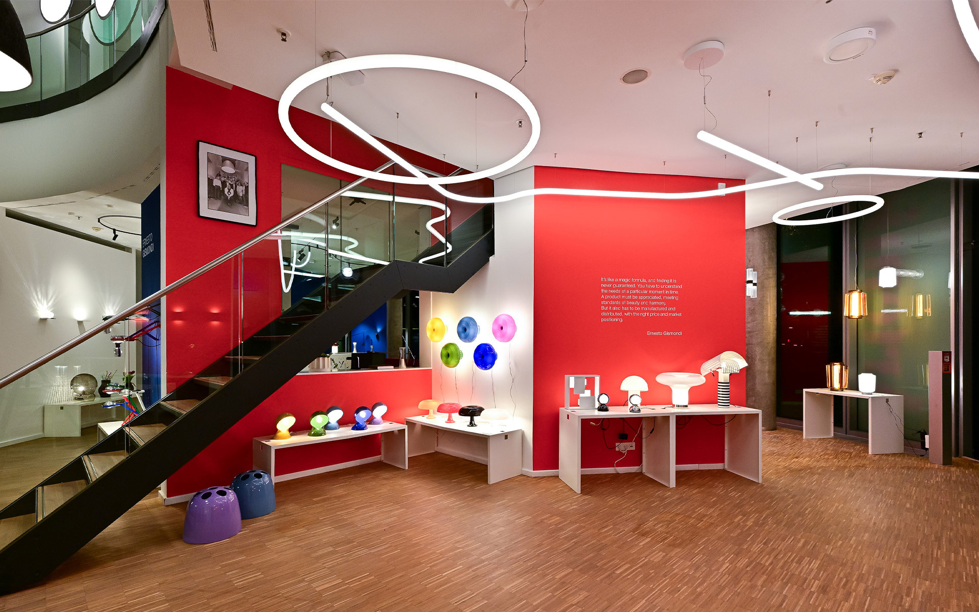 Artemide showroom with colorful table lamps, circular and wavy pendant lights on red walls, staircase, and lamp displays on white tables.