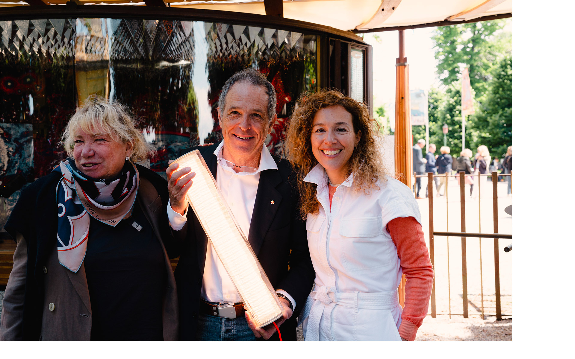 Three people smiling outdoors, center person holding an illuminated Artemide tubular LED lamp at an event.