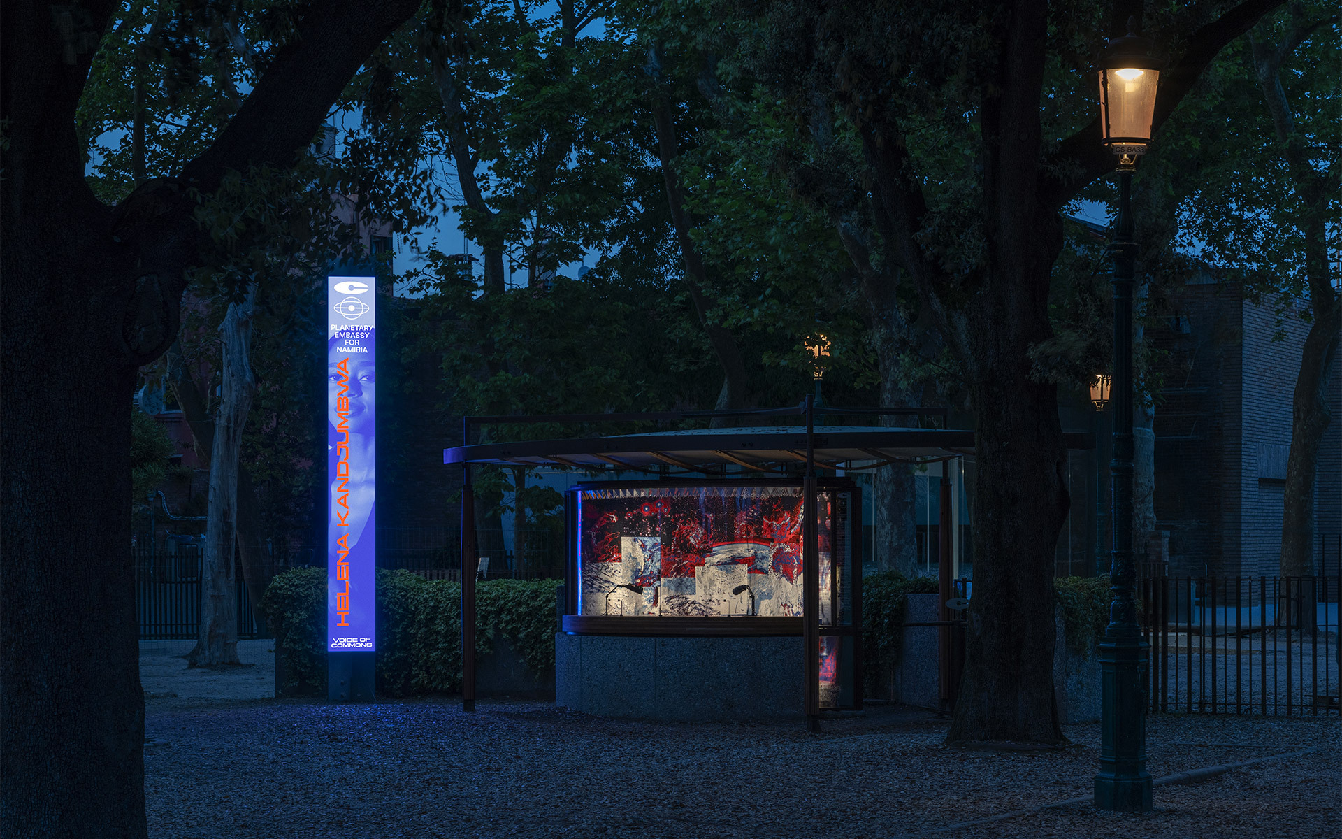 Illuminated outdoor kiosk and glowing vertical sign at dusk, surrounded by trees, with a classic street lamp casting warm light