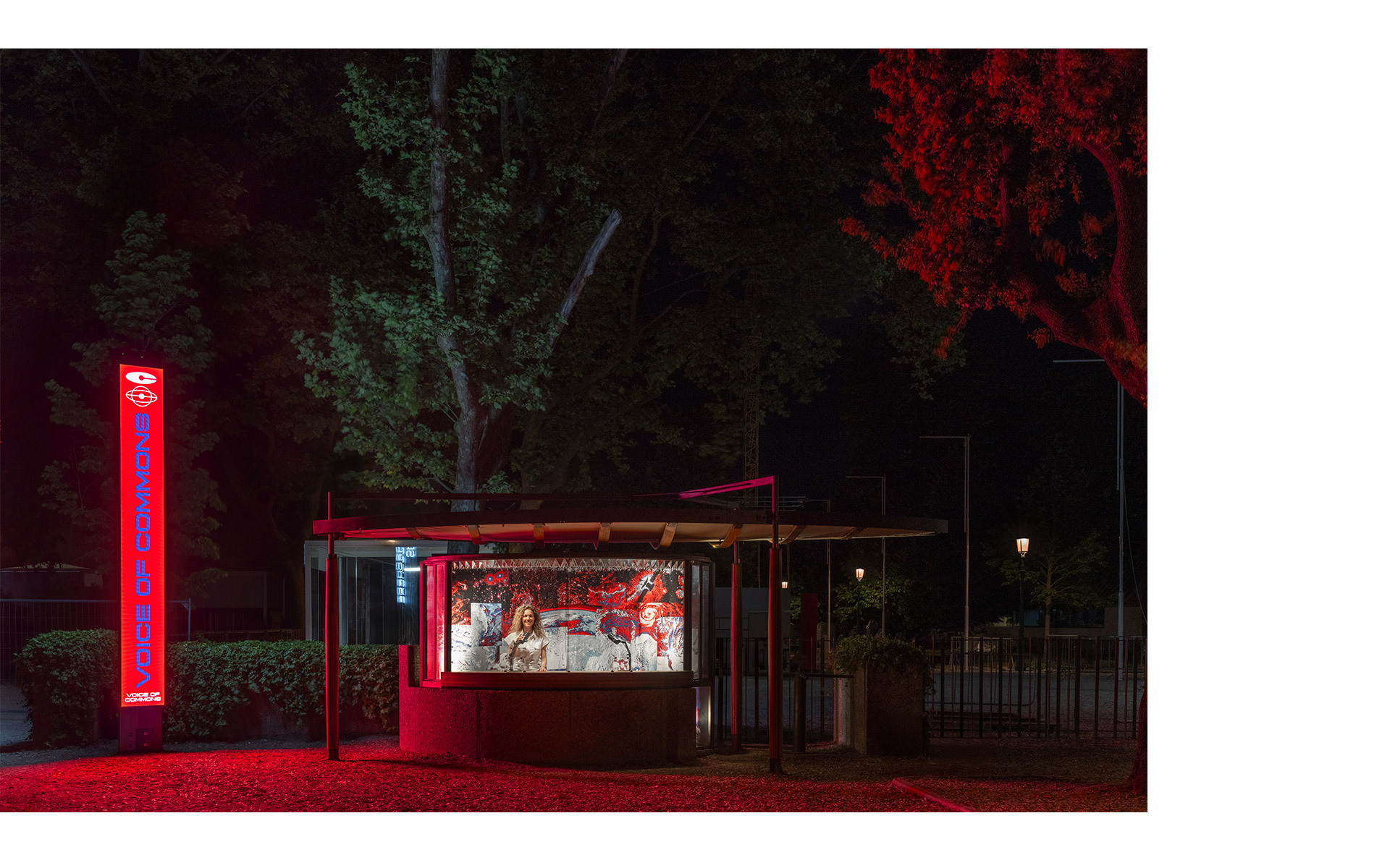 Red-lit outdoor kiosk at night with illuminated display window, neon signage, and trees dramatically lit in red light