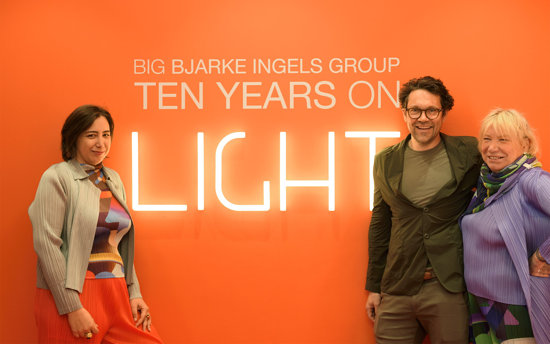 Three people posing in front of an orange wall with 'BIG Bjarke Ingels Group Ten Years on Light' illuminated sign.