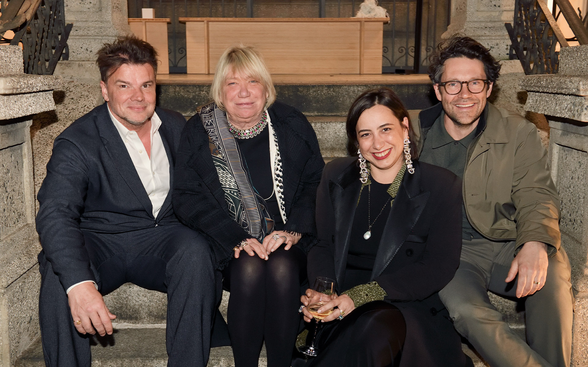 Four elegantly dressed people sitting together on stairs at a social event, smiling and holding drinks.