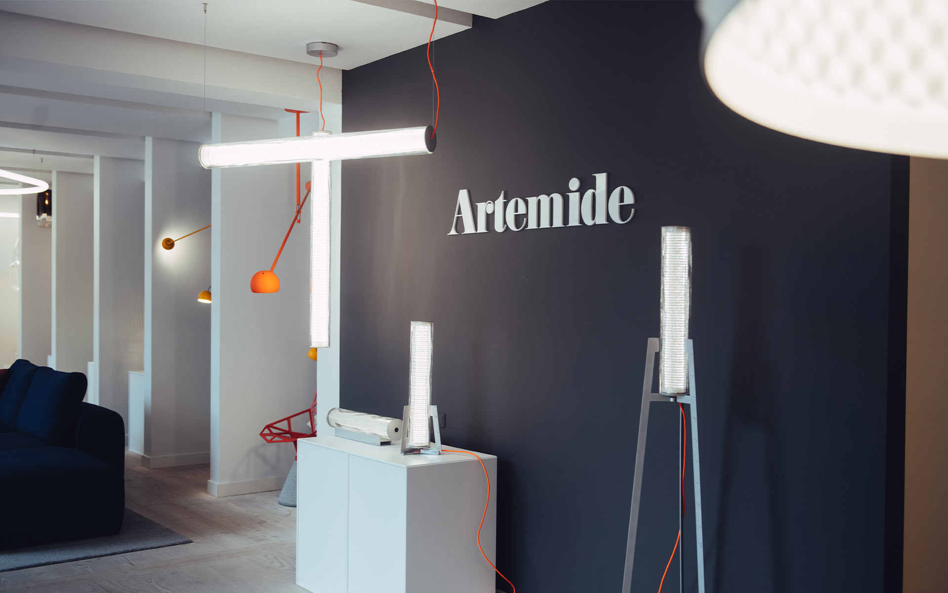 Artemide showroom displaying multiple luminaires: a horizontal LED tube pendant, orange wall lamps, and table lamps on white pedestals.