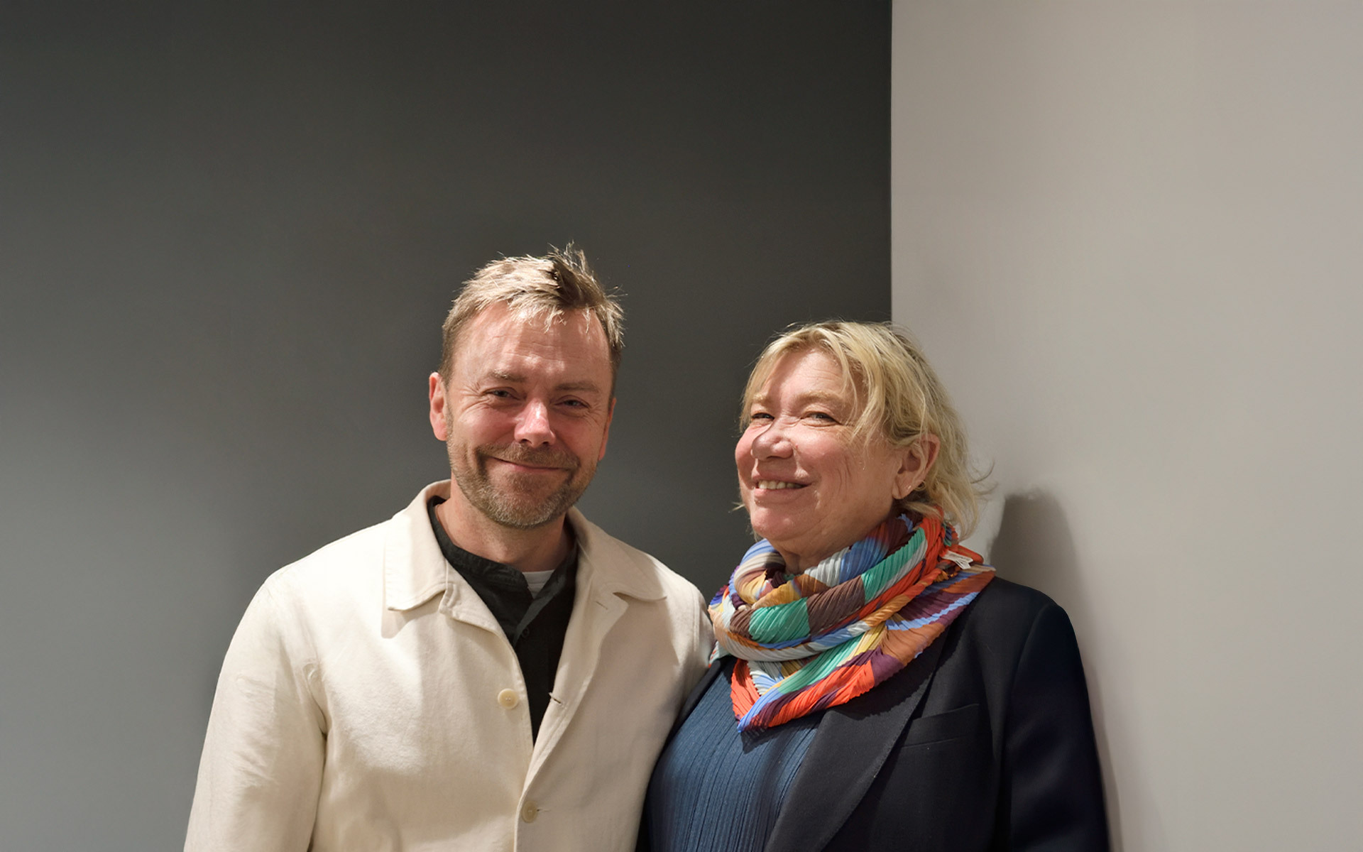 Two people smiling together: a man in a white jacket and a woman in a dark blazer with a colorful scarf.
