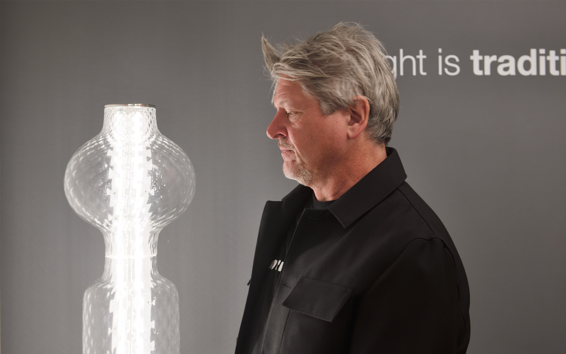 Designer in black standing beside a tall sculptural clear glass column lamp with stacked bulbous forms, in a showroom