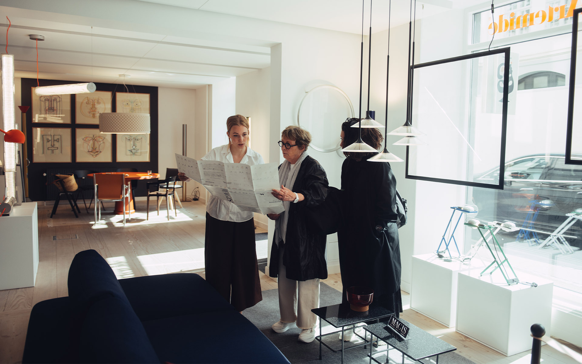 Three people consulting documents in an Artemide showroom, with pendant lamps overhead and lighting displays in the background.