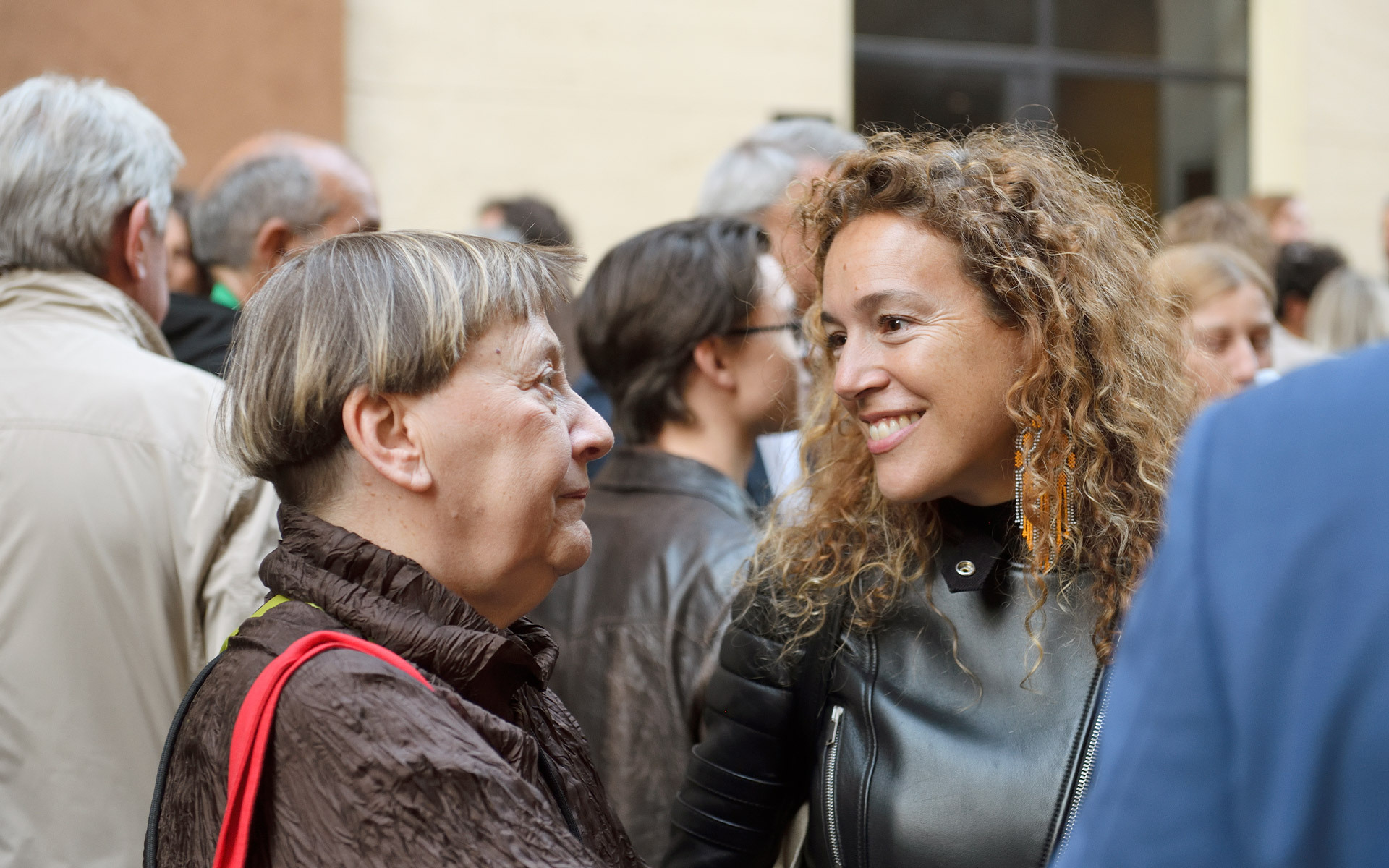 Two women engaged in conversation at a social gathering, one older with short gray hair, one younger with curly hair, smiling.