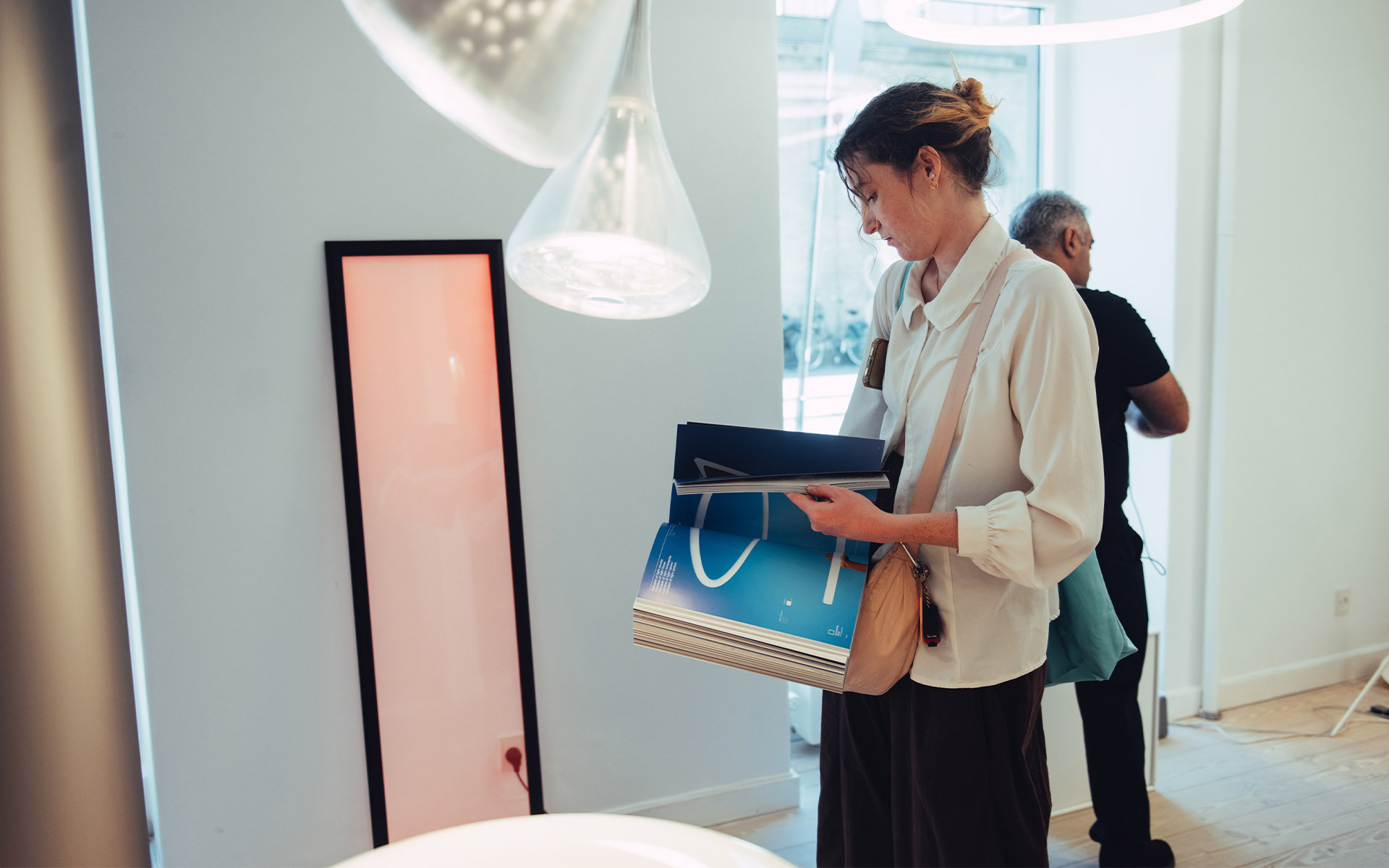 Woman browsing in a modern lighting showroom, holding a shopping bag, with a white pendant lamp and a pink floor lamp nearby.
