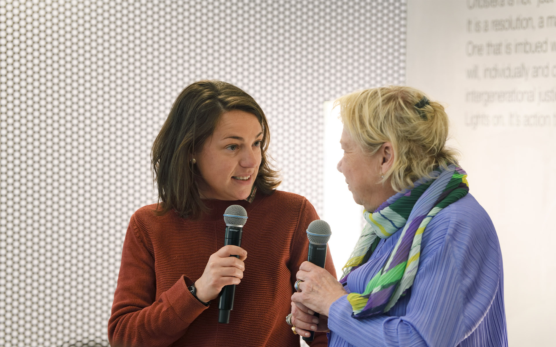 Two women holding microphones in conversation at an Artemide event, with a dotted wall panel and text display in the background.