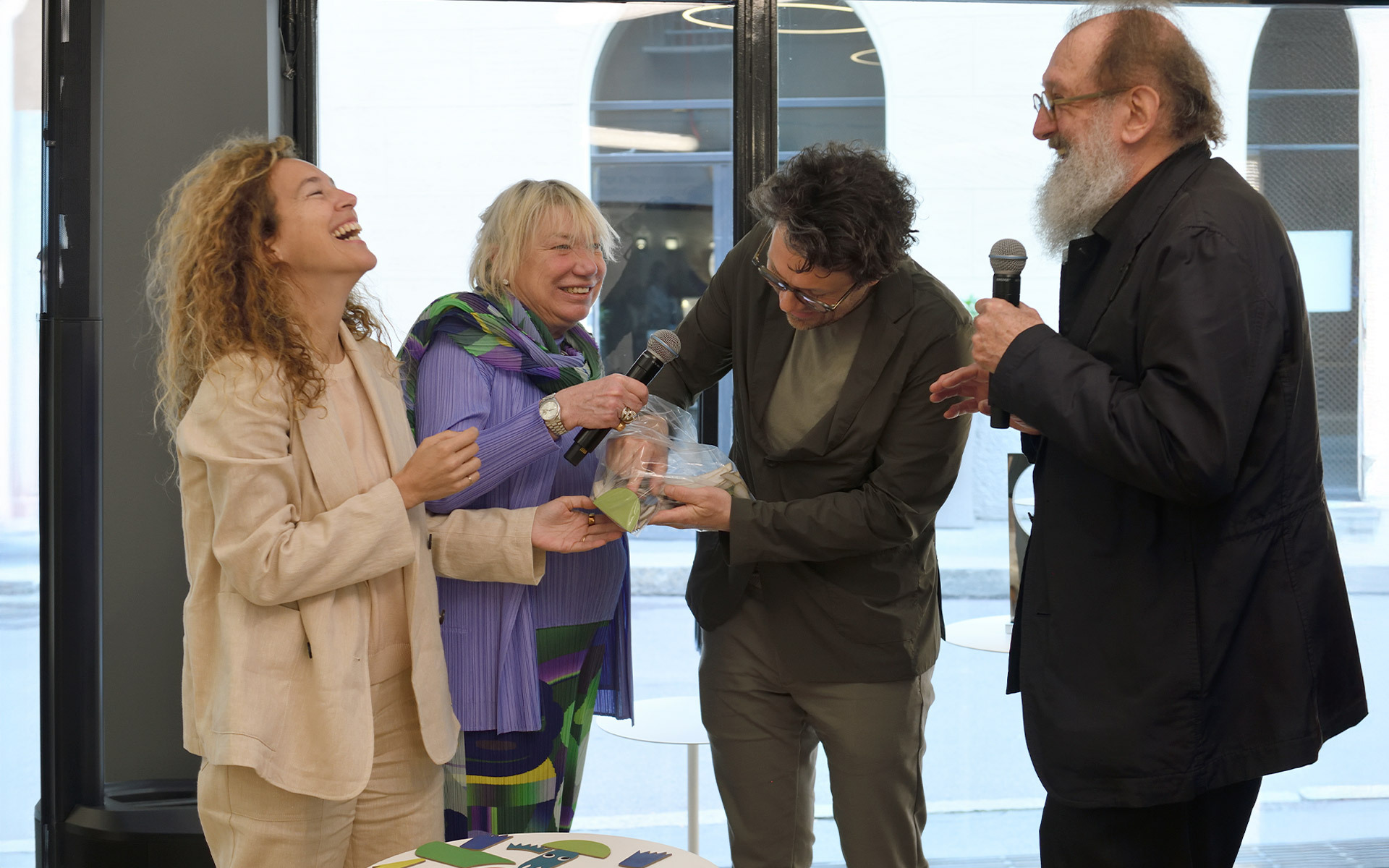 Four people laughing and talking at an event, one holding a microphone and another examining a small object.