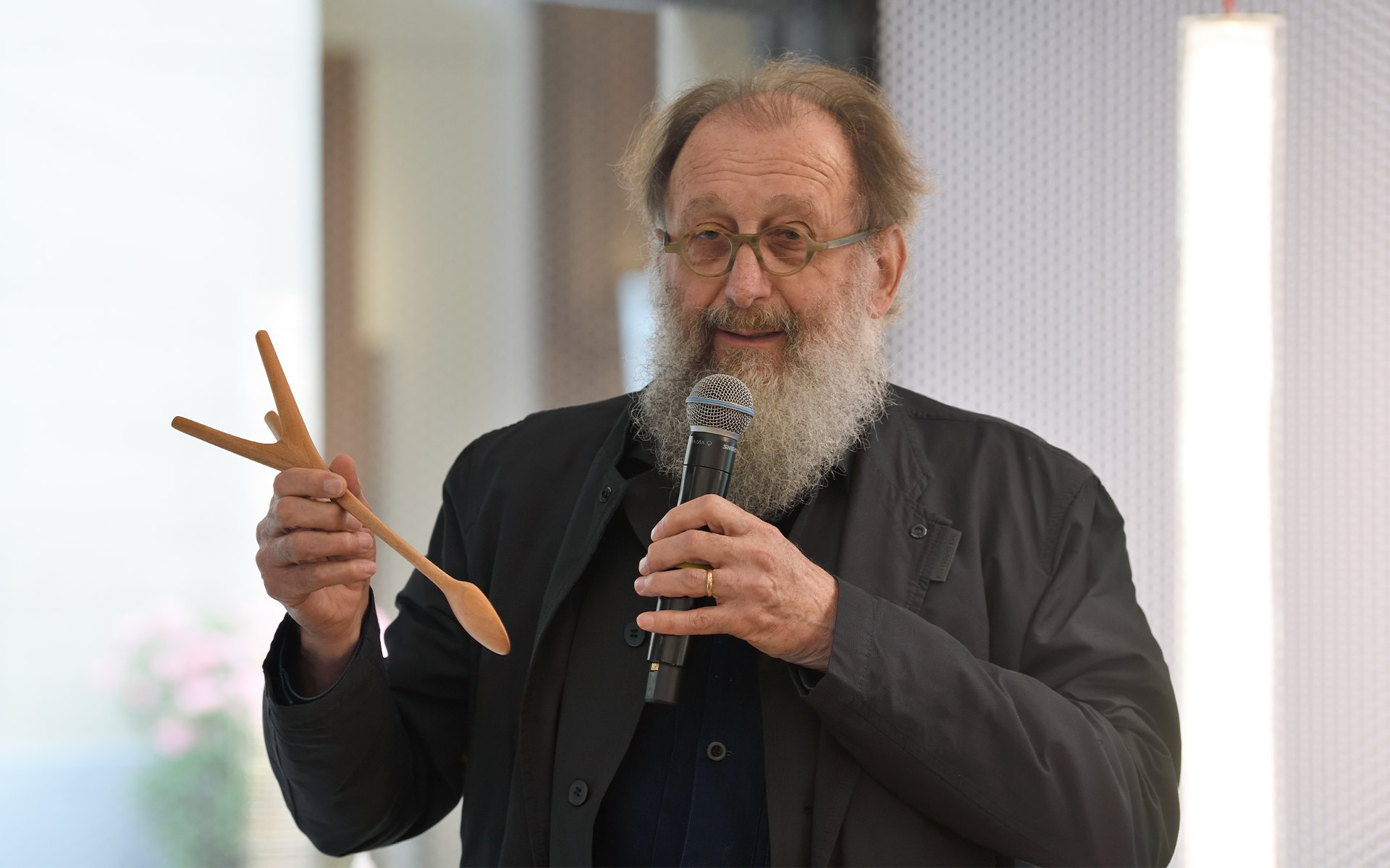 Bearded man in dark jacket speaks into microphone while holding a wooden spoon and fork-shaped object during a presentation