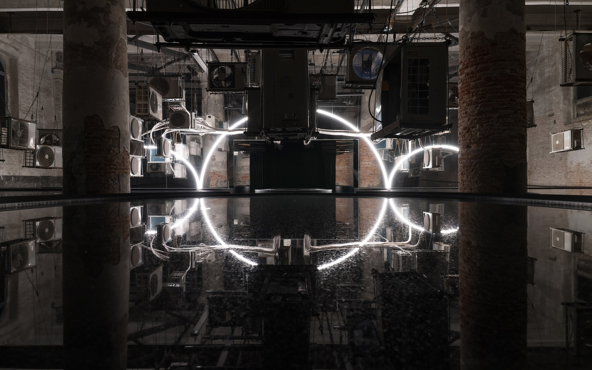 Glowing white tubular light installation forming a semicircle, reflected in a dark pool inside an industrial space with brick columns.