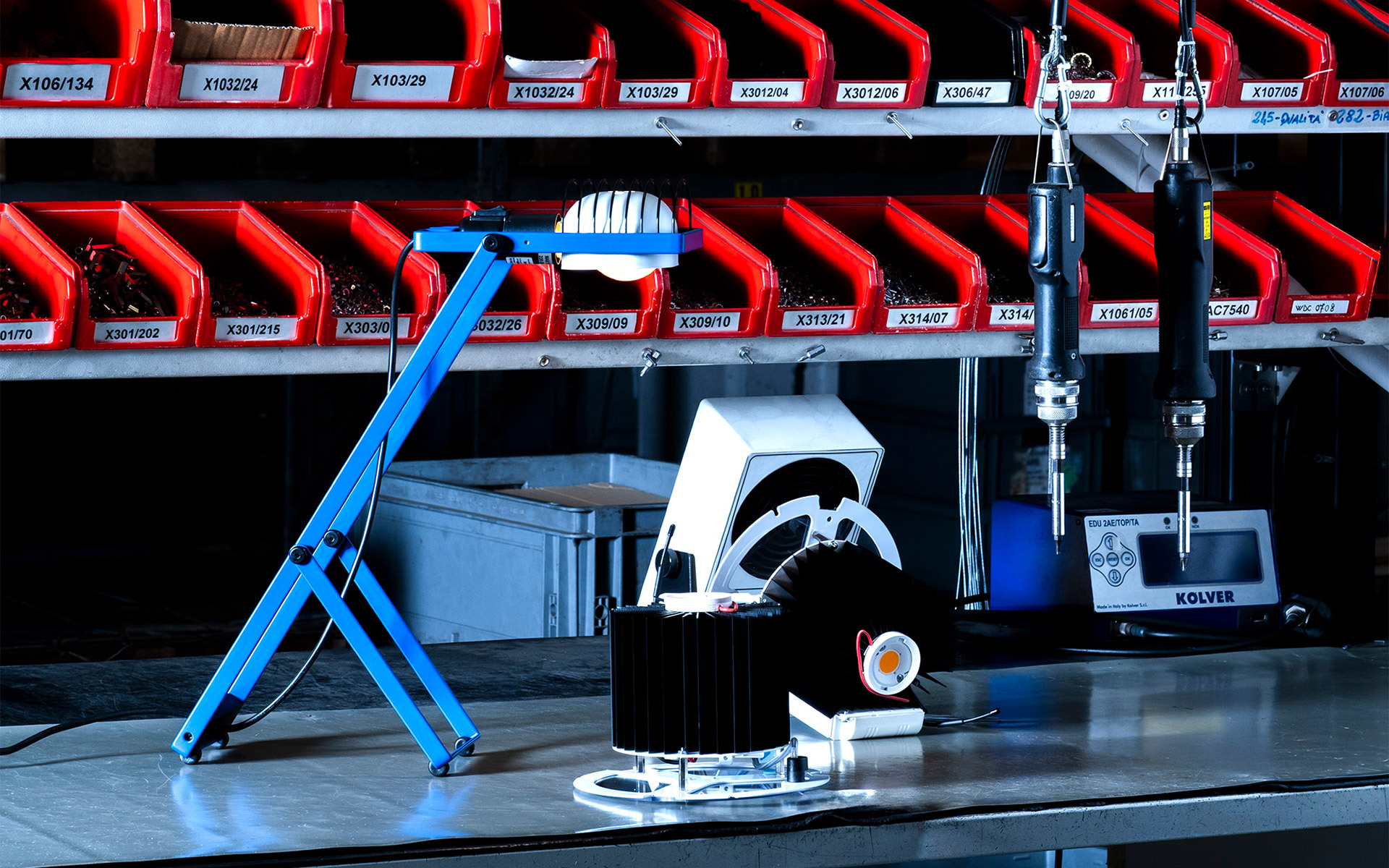 Blue adjustable desk lamp on a workbench in an industrial assembly area with red component storage bins in background