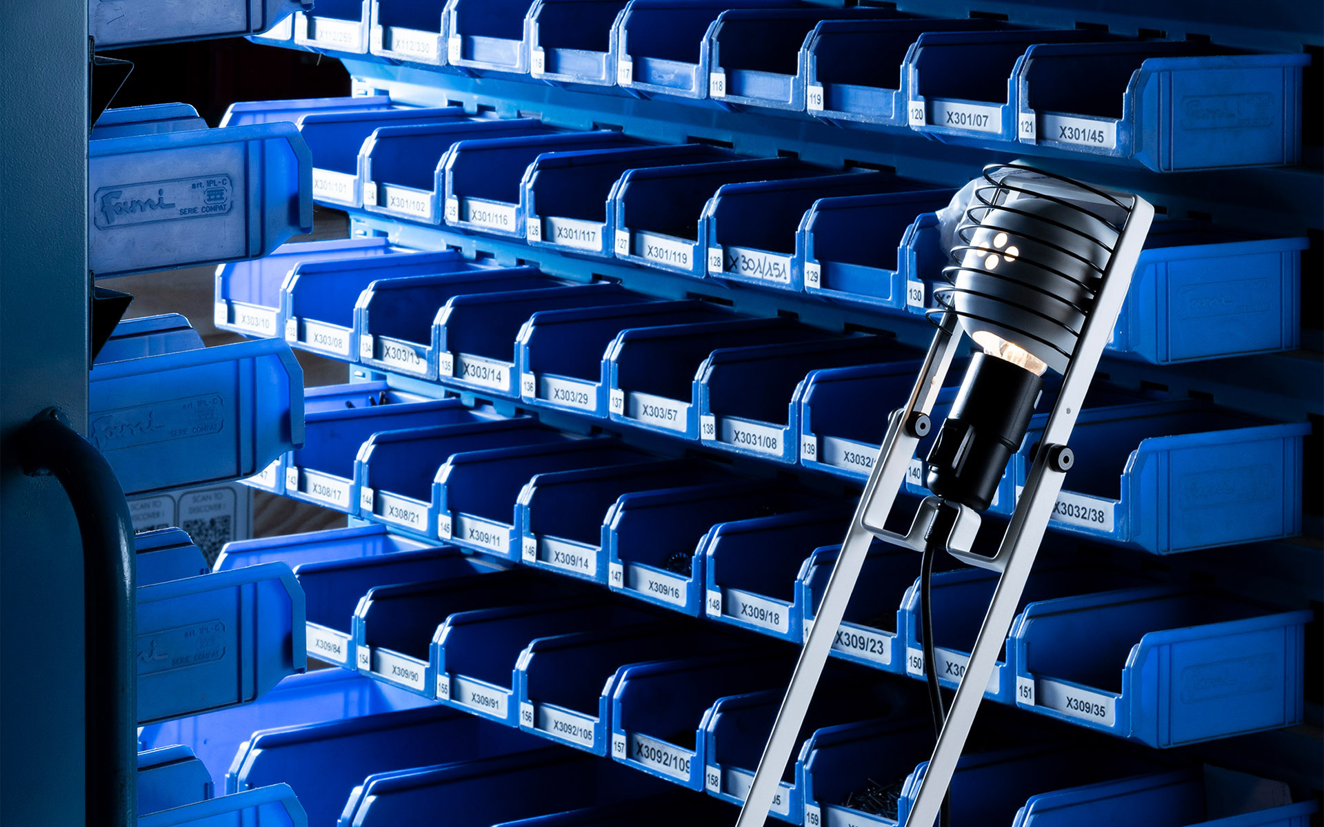 Artemide lamp with black cylindrical head on a ladder, illuminating blue industrial storage shelves with labeled bins