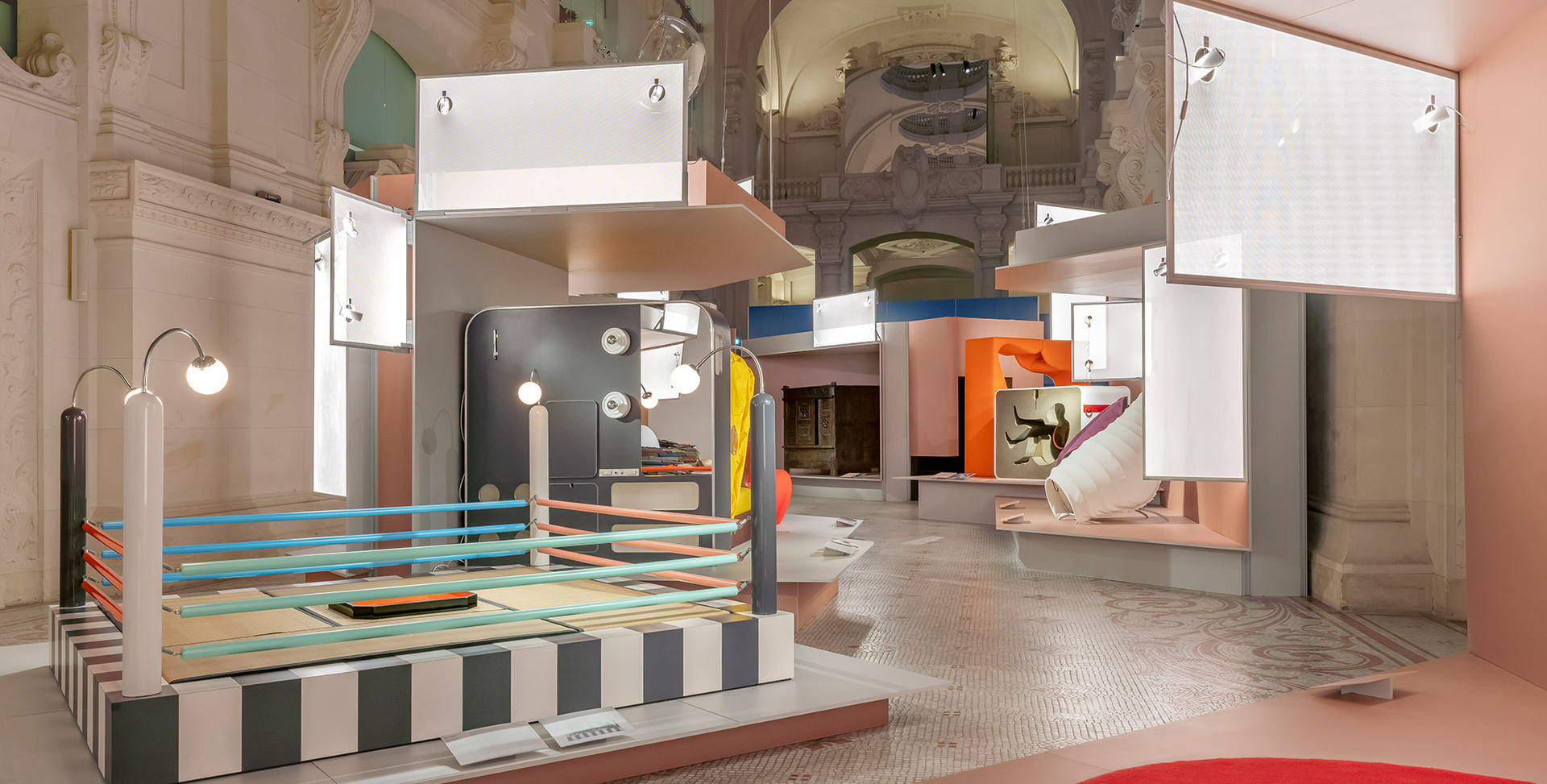 Colorful exhibition installation in a historic vaulted hall featuring geometric structures, suspended panels, and Artemide lighting fixtures.