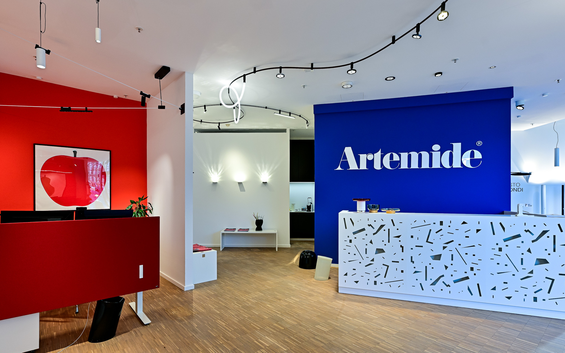 Artemide showroom entrance with branded blue reception desk, red accent wall, track lighting on ceiling, and wall-mounted lights.