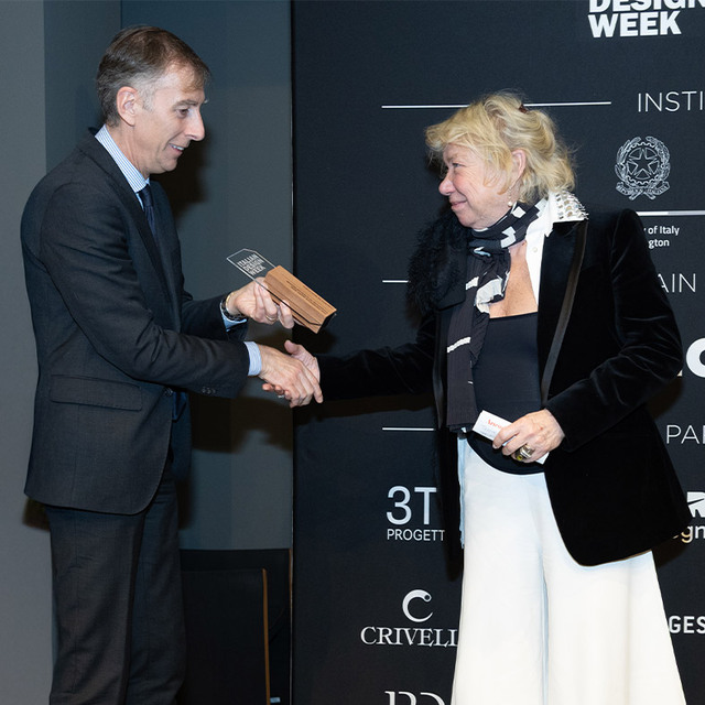 Two people shaking hands at a Design Week award ceremony, man in dark suit presenting a wooden trophy to a woman in black jacket.