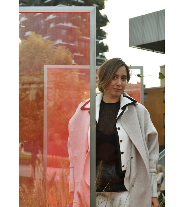 A woman in a light coat stands outdoors near a red translucent panel, with urban buildings and greenery in the background.