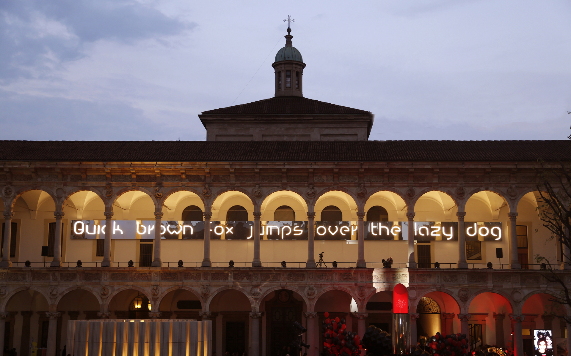Illuminated text 'Quick brown fox jumps over the lazy dog' projected on a classical Italian building arcade at dusk