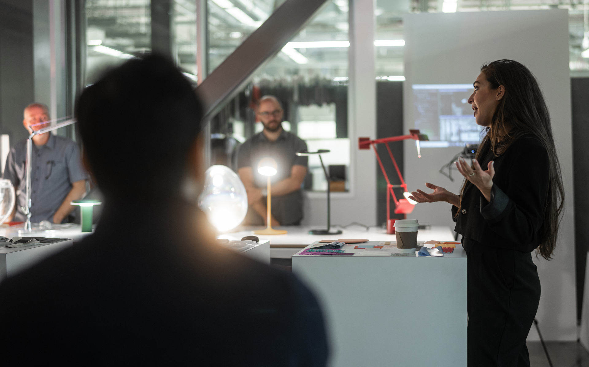 Presenter speaks to colleagues in a design studio with Artemide table lamps, including a red Tolomeo and a bright globe lamp on the desk.