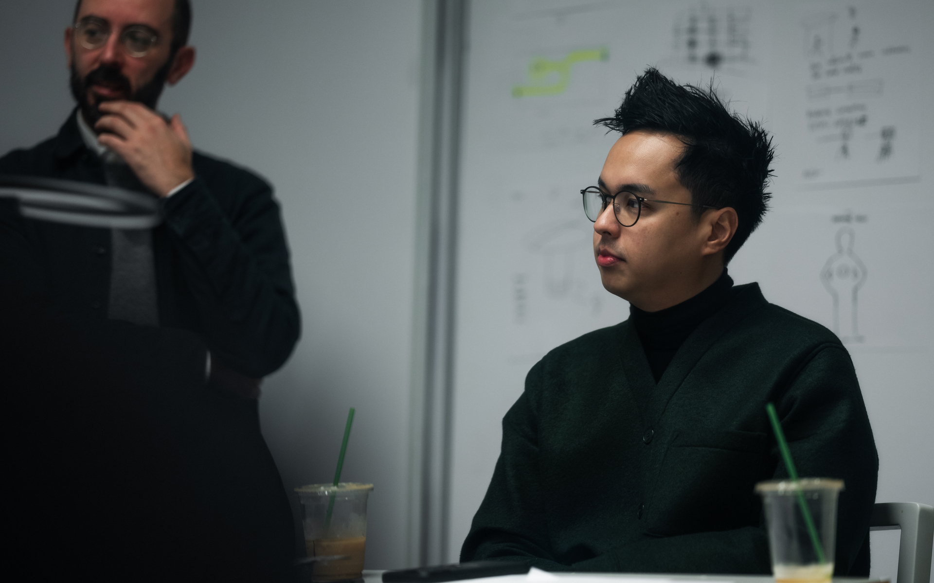 Two people in a design meeting: one standing thoughtfully, one seated wearing glasses, with sketches on a whiteboard behind them.