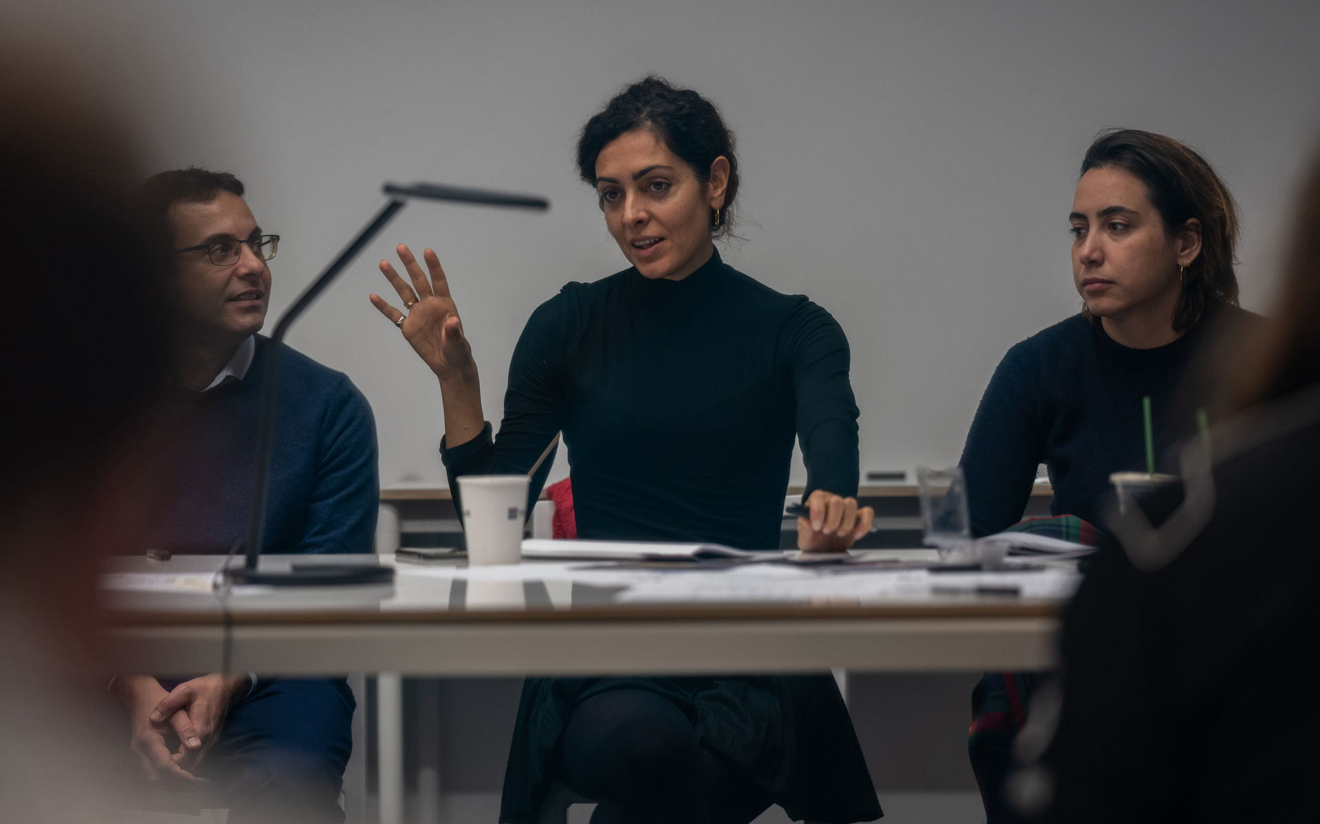 Three people at a meeting table, woman in center gesturing while speaking, Artemide desk lamp visible in background