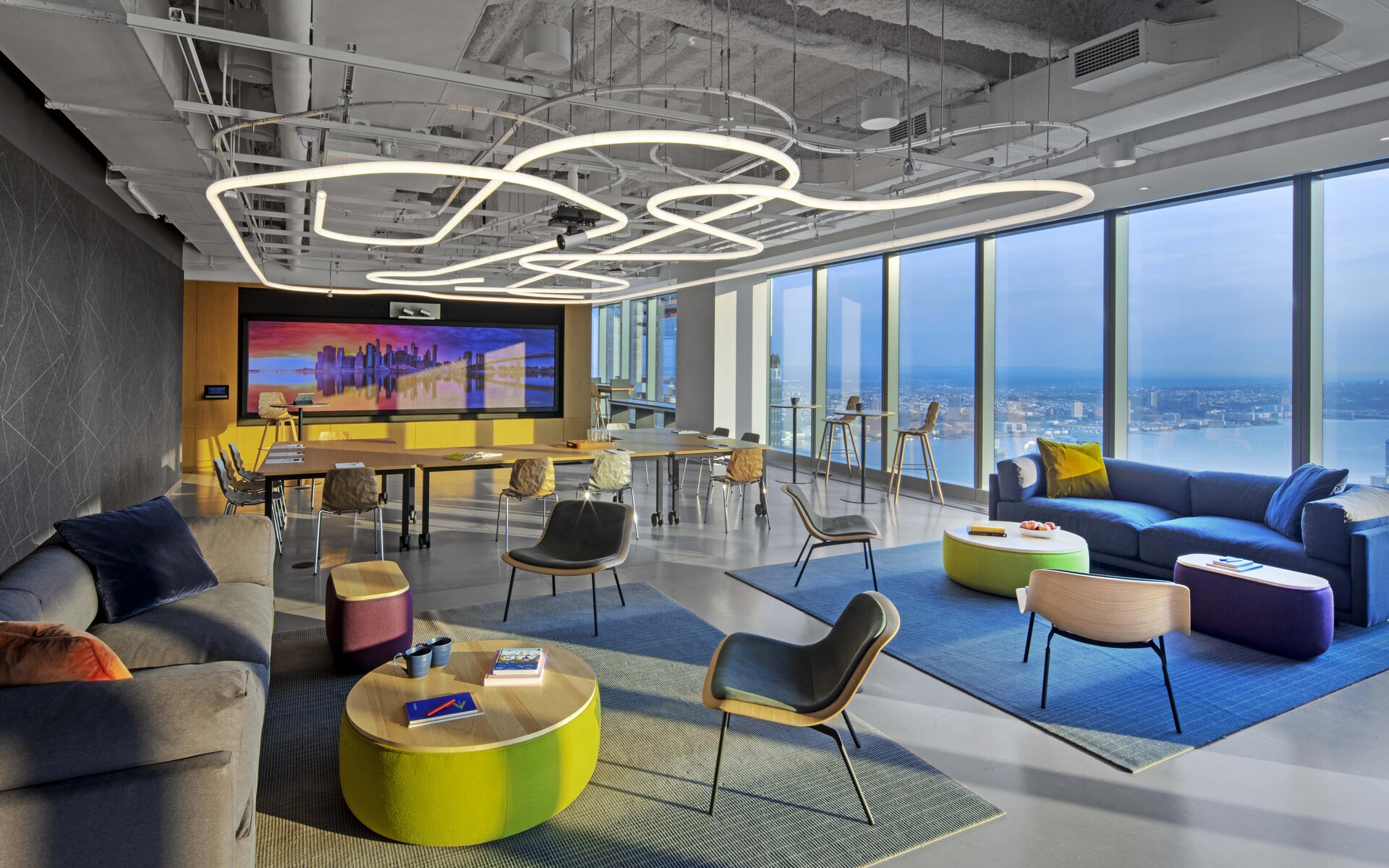 Modern office lounge with Artemide pendant lights forming interconnected rings overhead, colorful furniture, and panoramic city views.