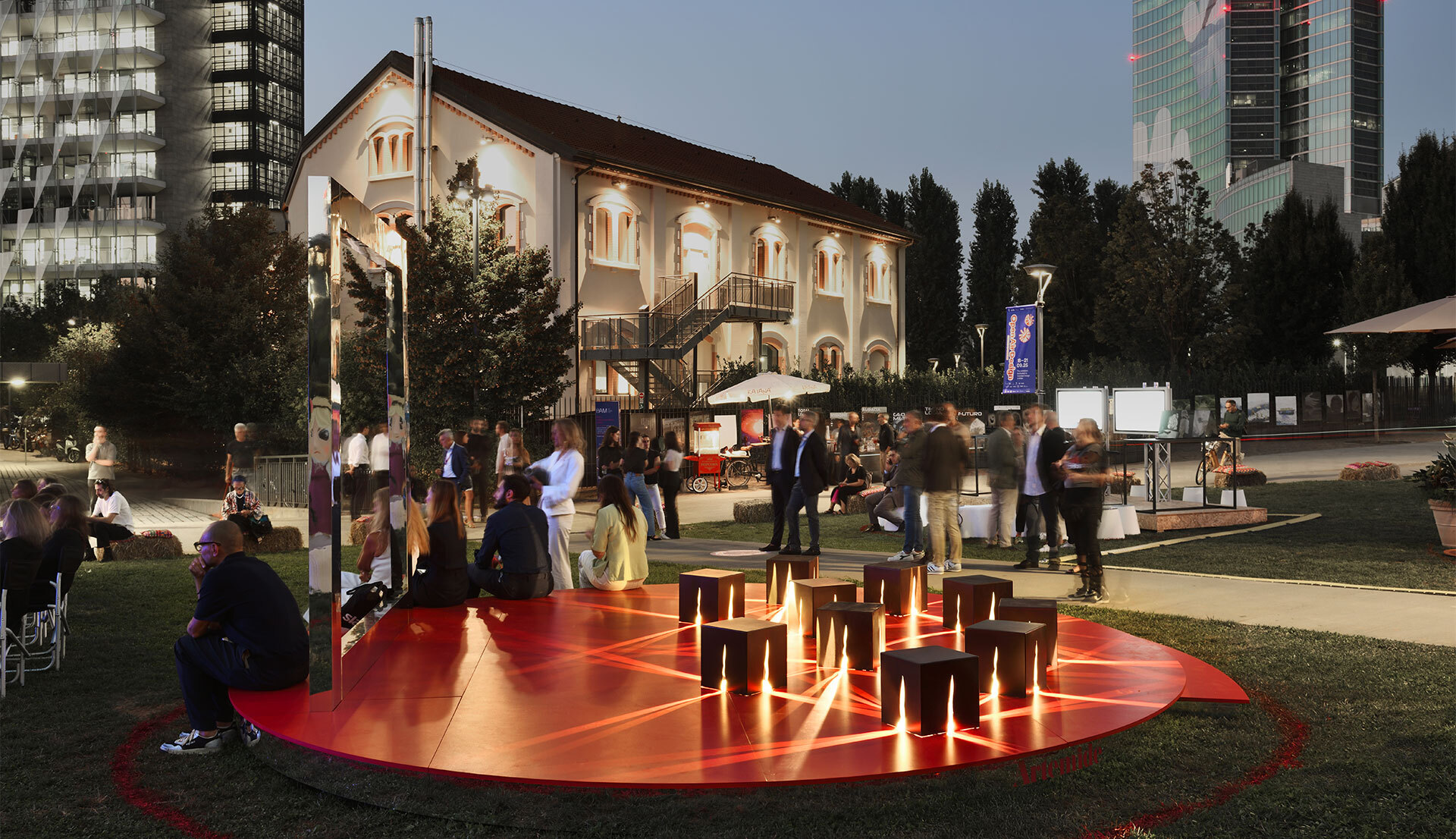 Outdoor evening event with people gathered around a glowing red circular platform with wooden stools, lit from below, urban setting.