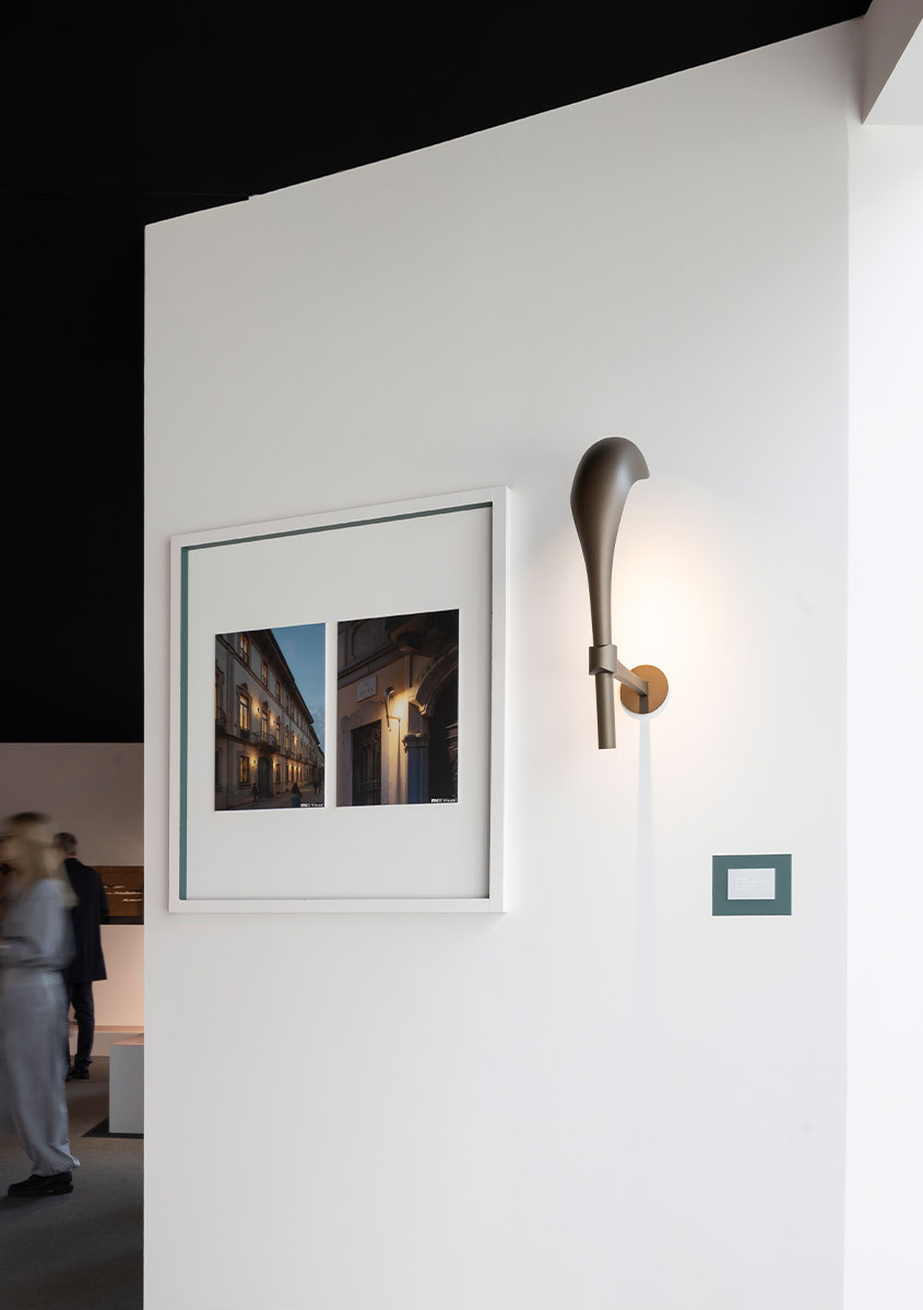 Artemide wall sconce with elongated dark cone shade and brass mount, displayed on white wall beside framed architectural photography.