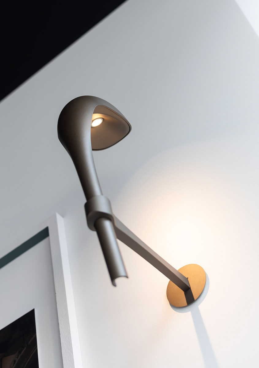 Artemide wall lamp with elongated dark arm, upward-facing cone shade with LED light, mounted on a gold circular wall base