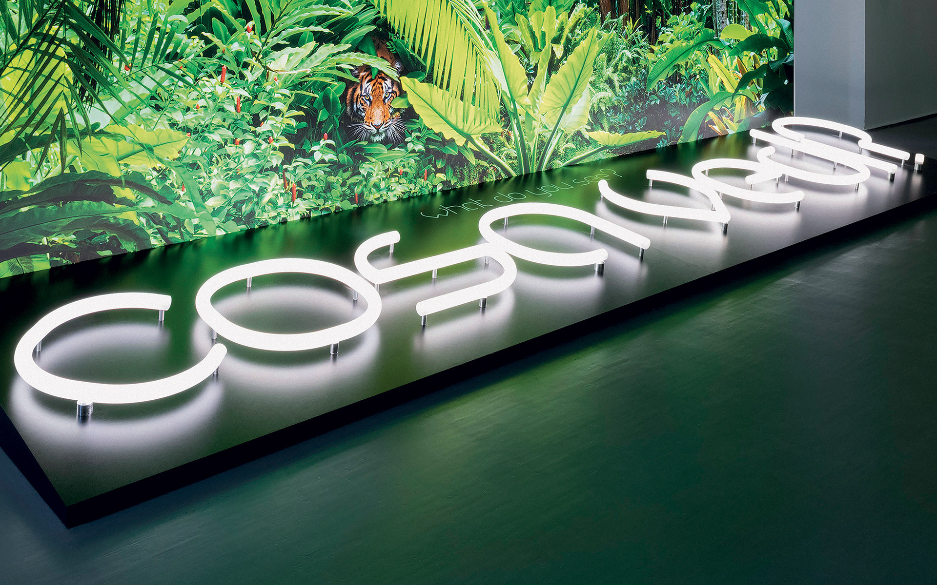 Illuminated white neon letters spelling 'cosmo' on a dark base, set against a lush tropical jungle backdrop with a tiger.