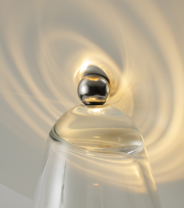 Close-up of a clear glass lamp top with a chrome spherical finial, casting concentric light ripples on the ceiling above