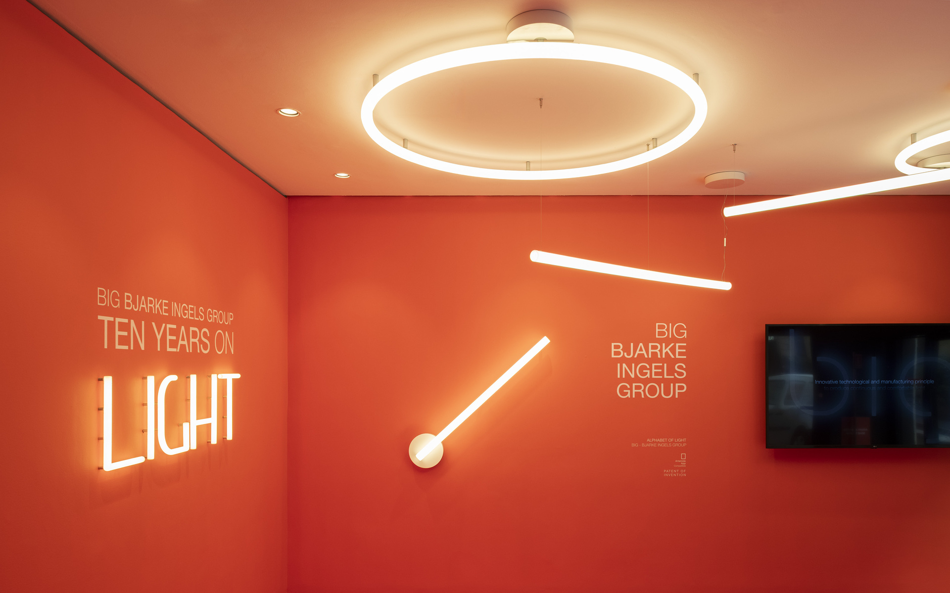 BIG Bjarke Ingels Group exhibition with Artemide neon and circular LED ceiling lights on orange walls, titled 'Ten Years on Light'