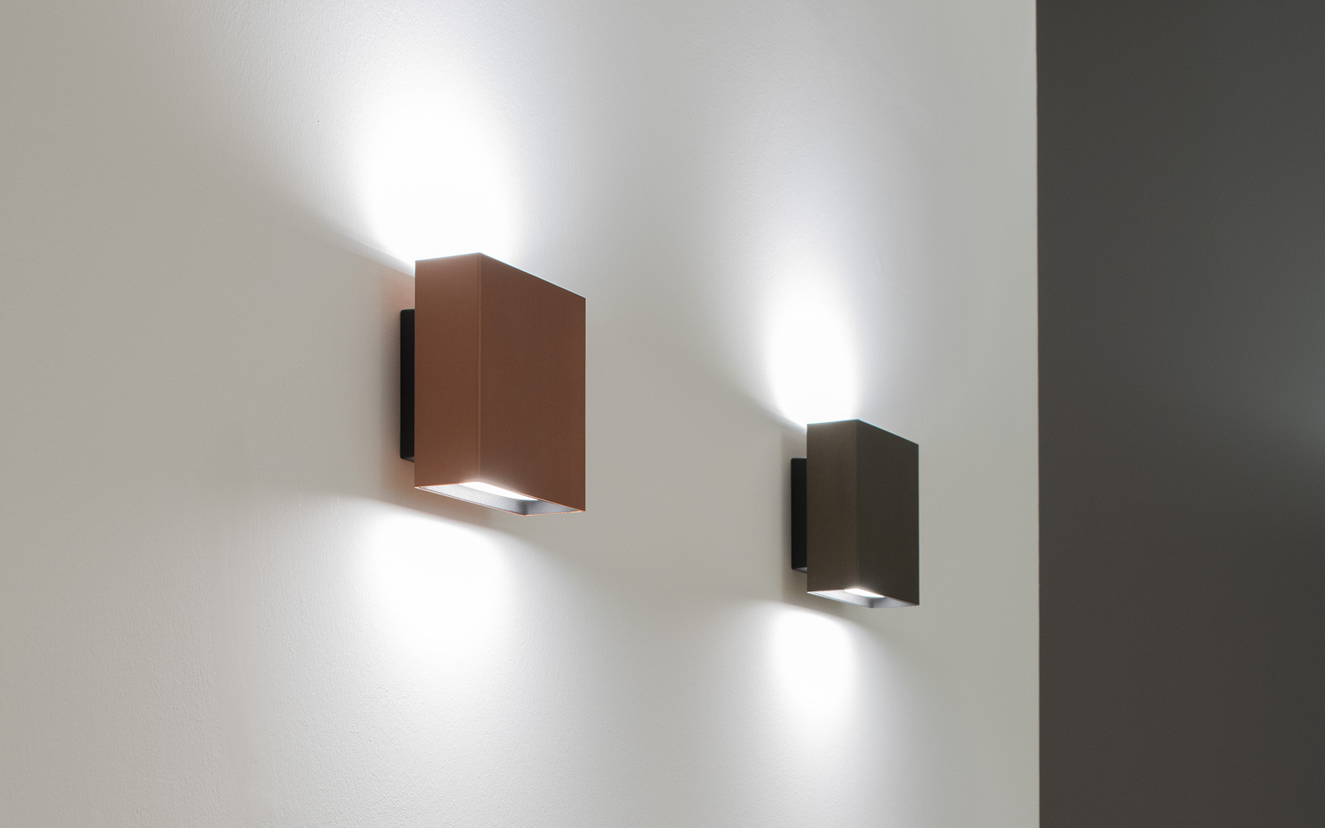 Two square wall sconces in copper and dark bronze finishes, emitting upward and downward light beams against a white wall.