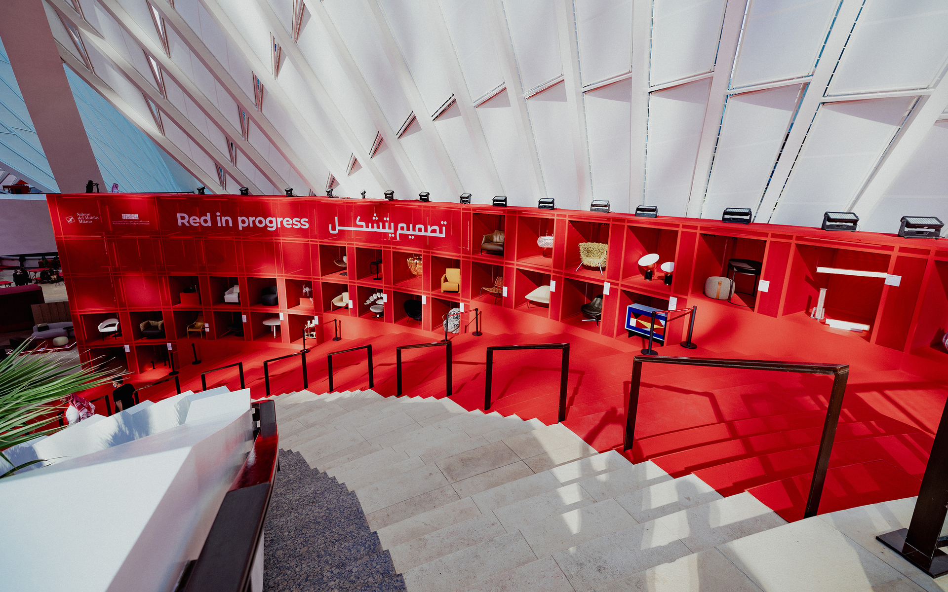Red in Progress exhibition: bold red display wall with shelved design objects and lamps, Arabic and English signage, viewed from stairs.
