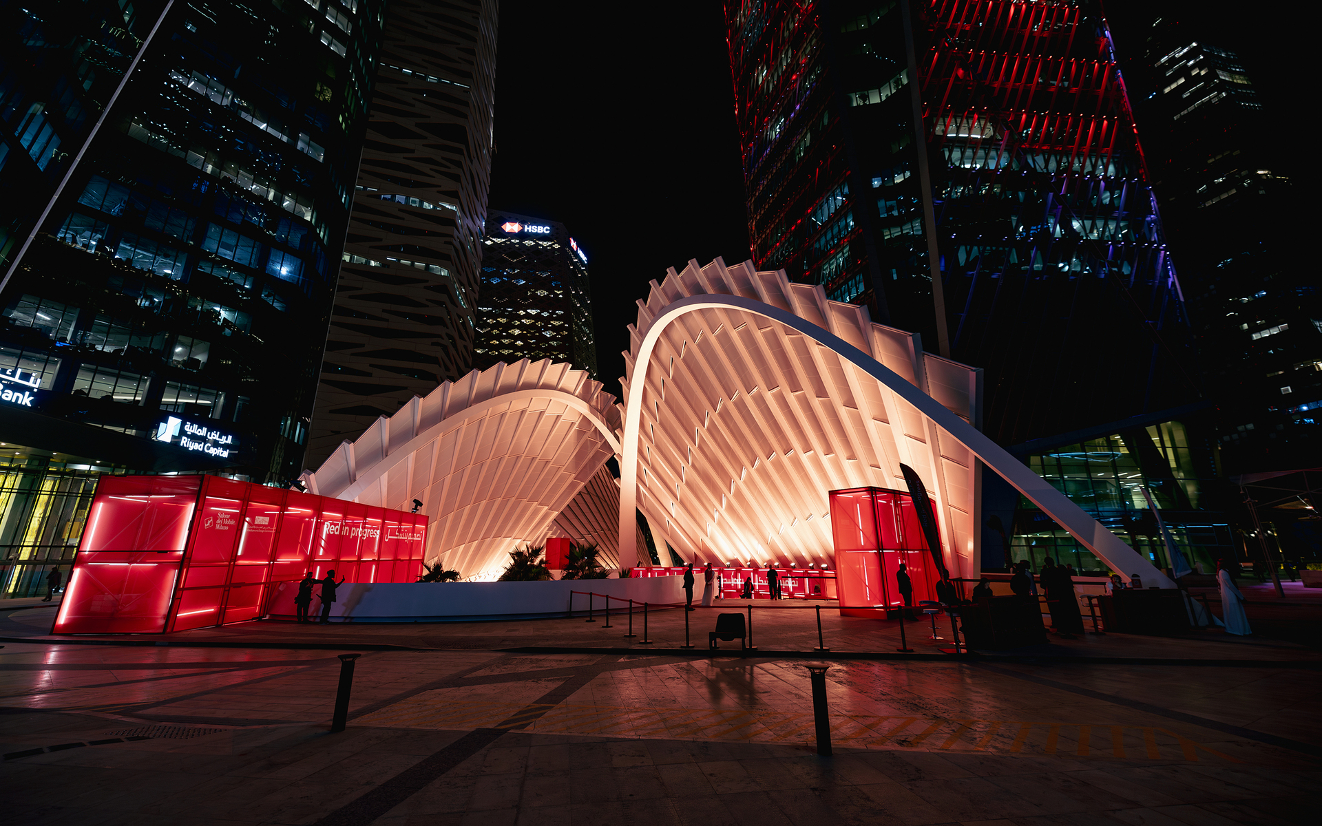 Illuminated white architectural pavilion with arched ribbed canopy and red-lit side panels, set against a night cityscape