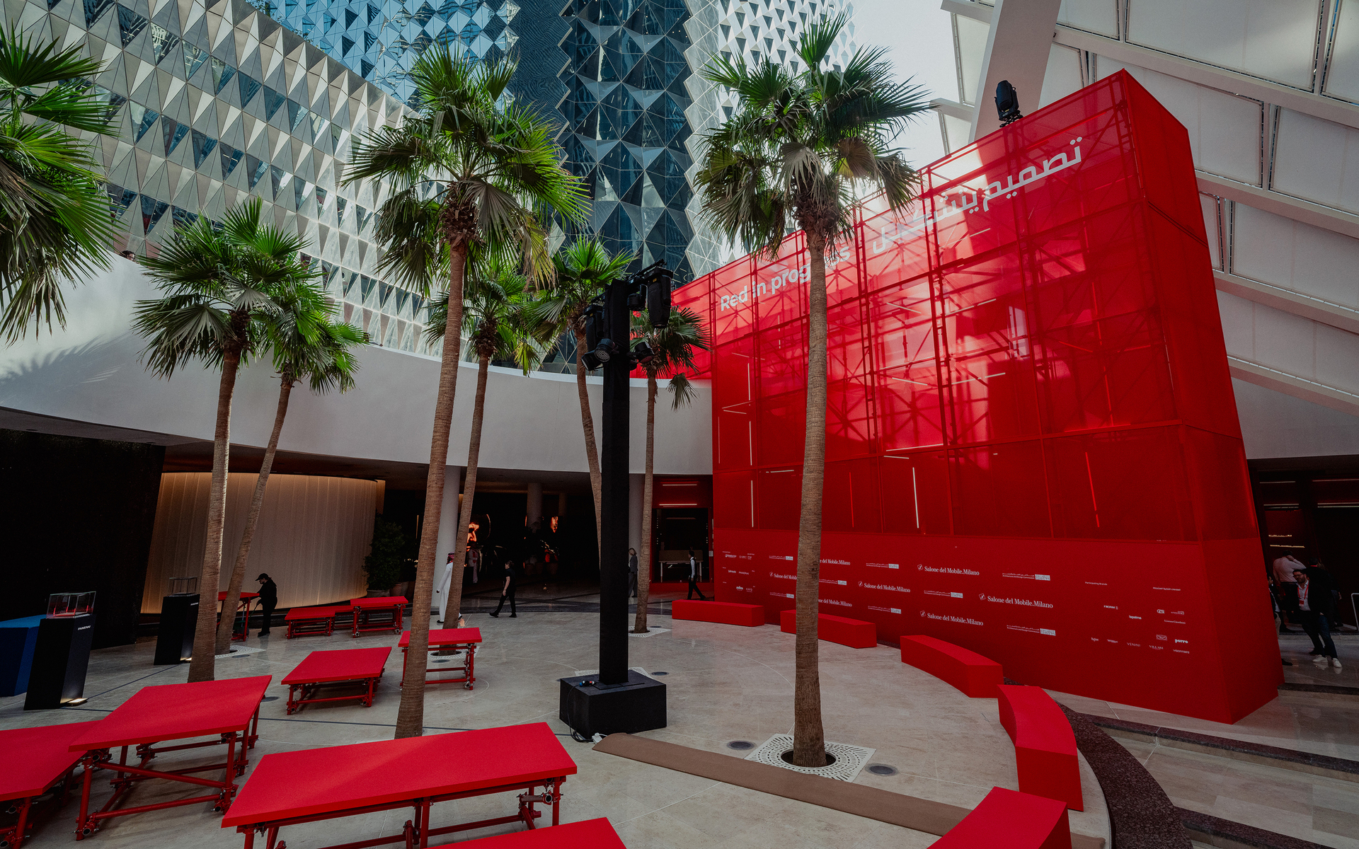 Outdoor plaza with red benches, palm trees, and a large red translucent structure with Arabic script, flanked by modern buildings.
