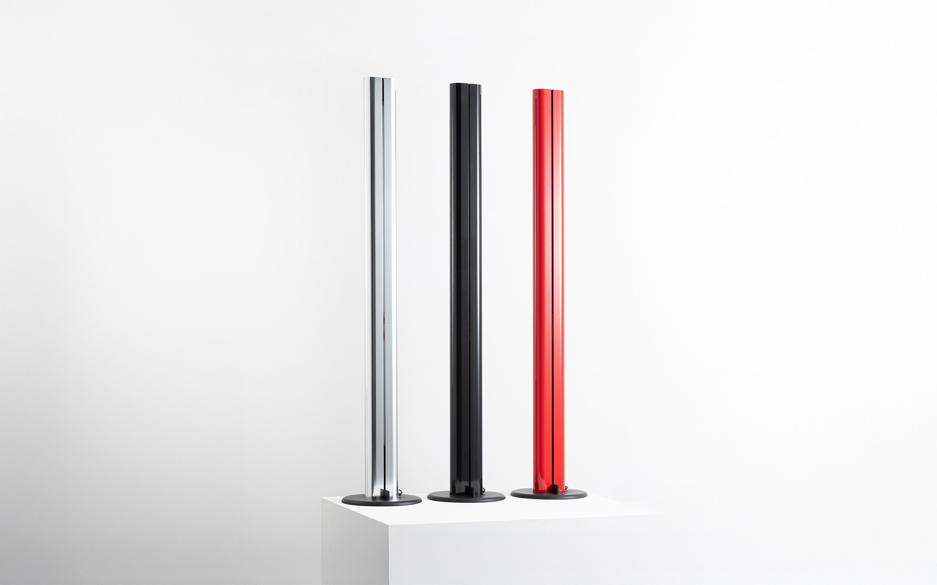 Three slim floor lamps on round bases in silver, black, and red, displayed on a white pedestal against a neutral background.