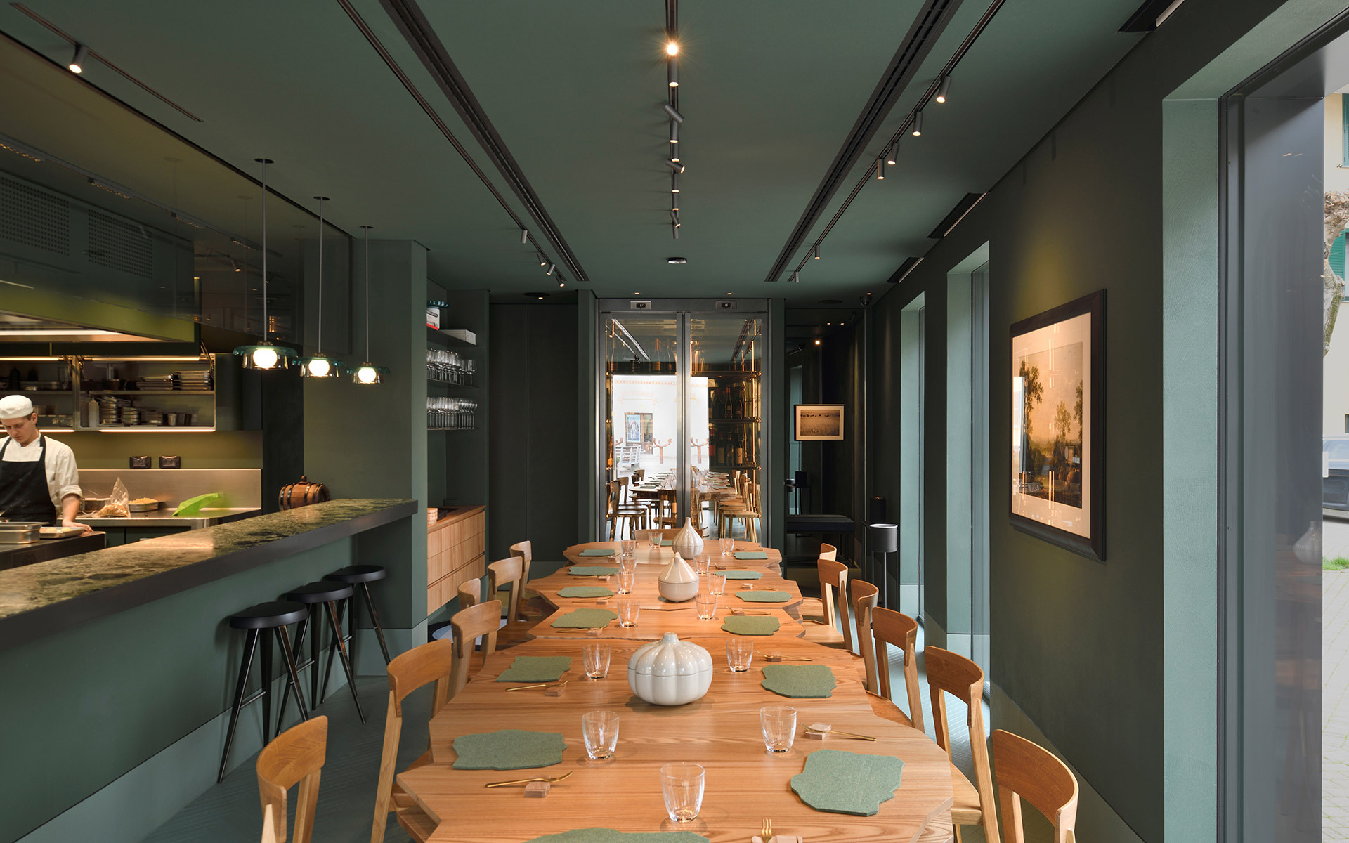 Restaurant dining room with Artemide track lighting on teal ceiling, long wooden table set for dinner, open kitchen with pendant lights