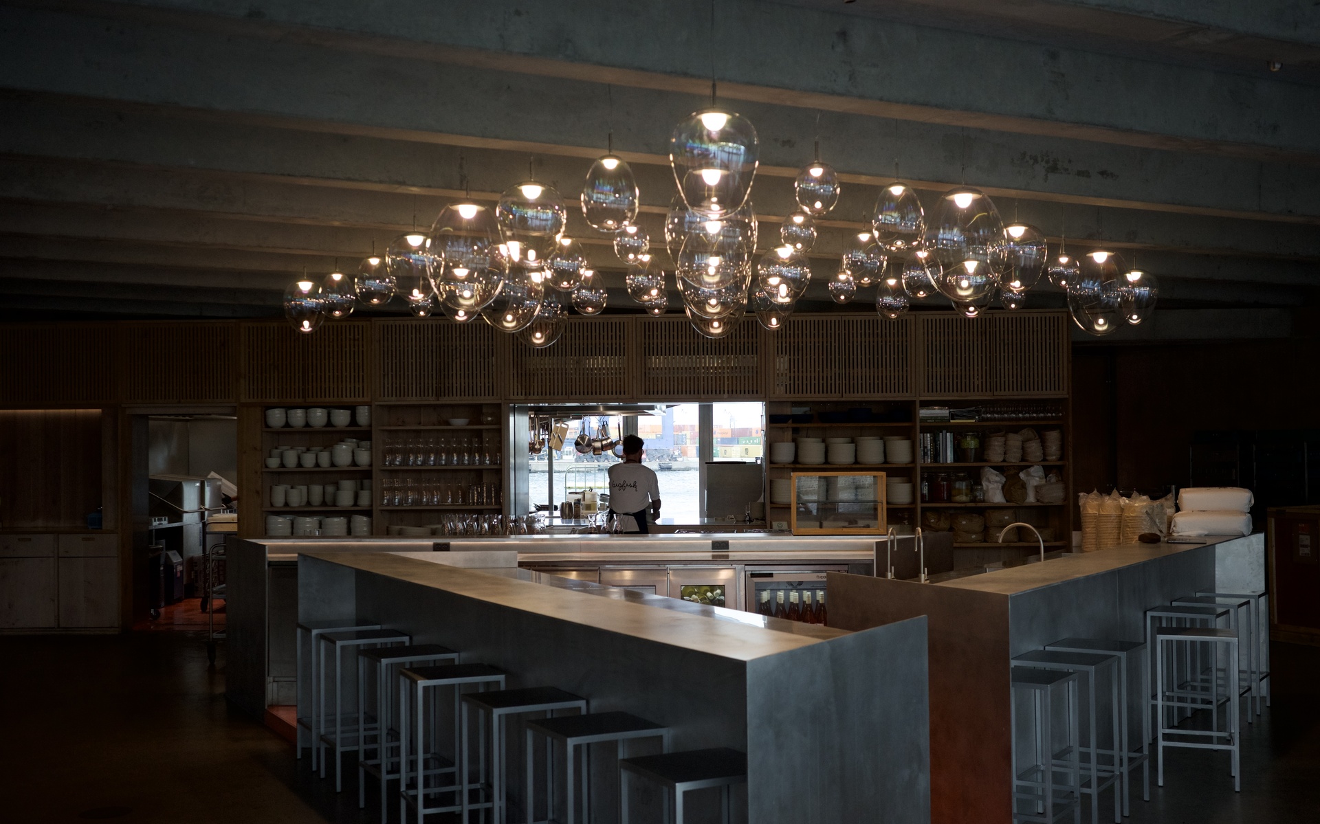 Restaurant bar with large cluster chandelier of glass globe pendant lights illuminating a concrete island counter with metal stools.