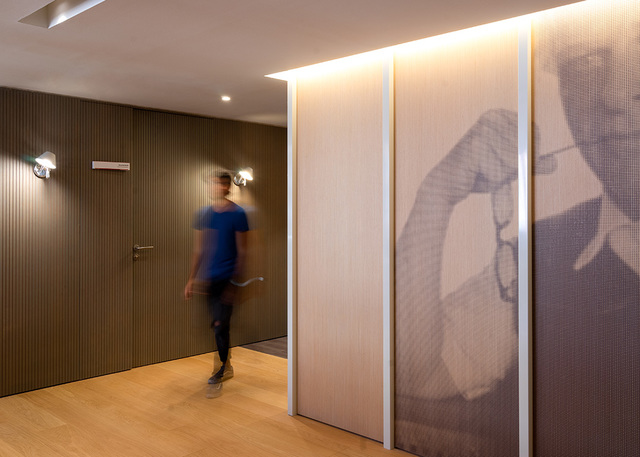 Modern corridor with warm LED cove lighting, wall-mounted spotlights, and a person walking past a decorative printed panel.
